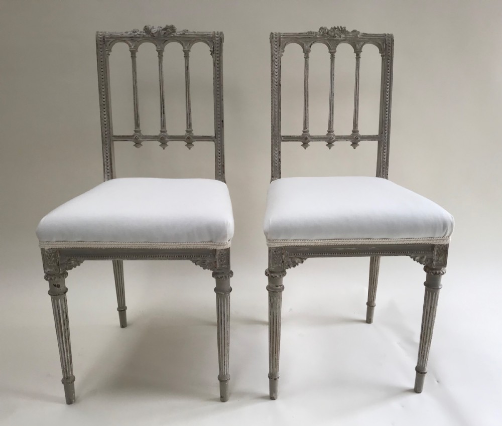 A pair of French Louis XVI style side chairs featuring distressed grey painted wooden frames, carved arcade backrests, and neutral white upholstered seats.