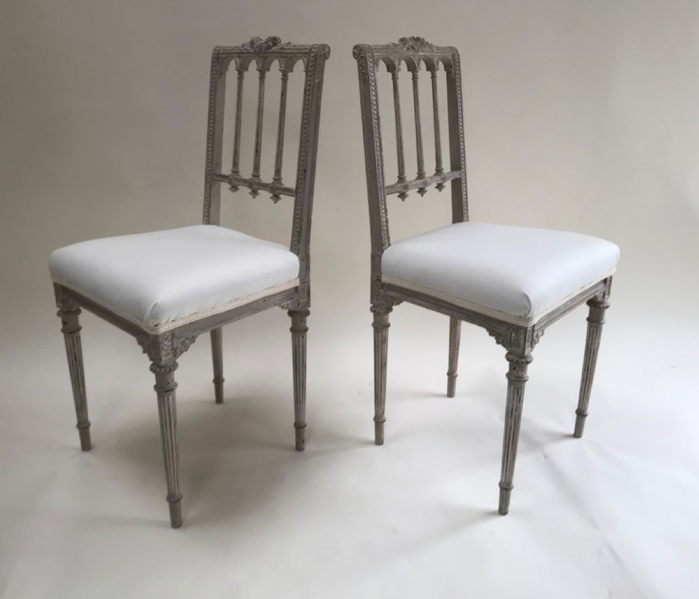 A pair of French Louis XVI style side chairs featuring distressed grey painted wooden frames, carved arcade backrests, and neutral white upholstered seats. View IV