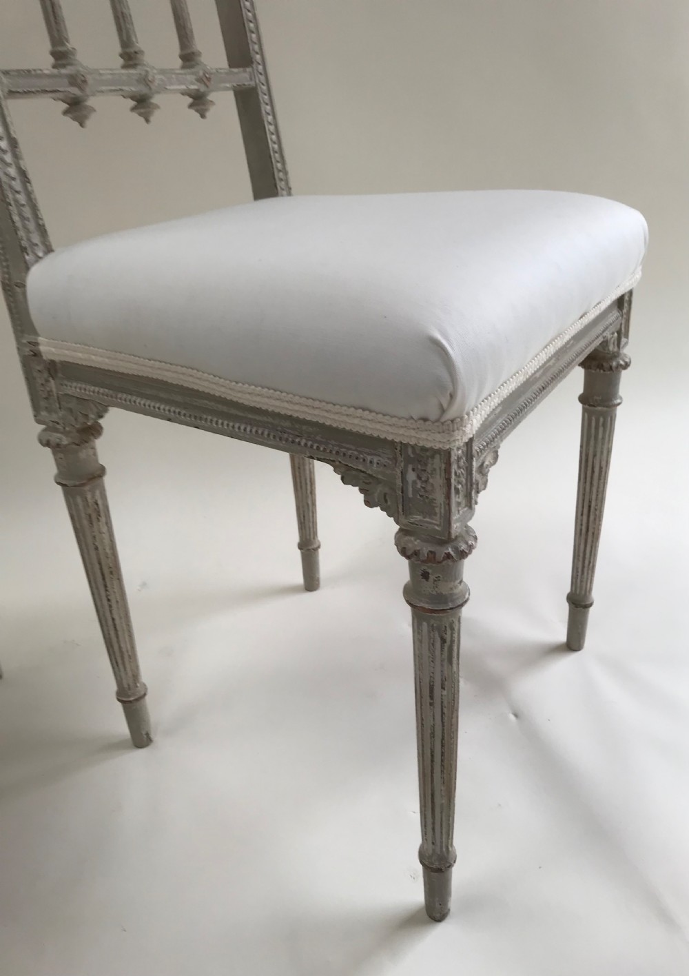 A pair of French Louis XVI style side chairs featuring distressed grey painted wooden frames, carved arcade backrests, and neutral white upholstered seats. View VI