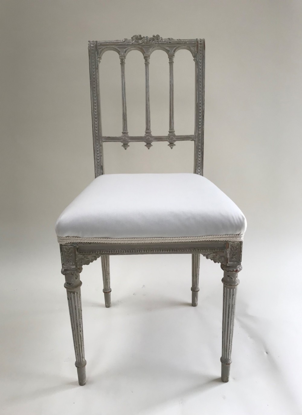 A pair of French Louis XVI style side chairs featuring distressed grey painted wooden frames, carved arcade backrests, and neutral white upholstered seats. View VII