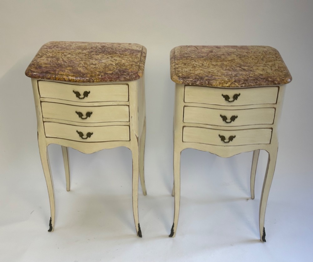 A pair of cream-painted French bedside tables featuring variegated marble tops, three drawers with brass pulls, and slender cabriole legs.