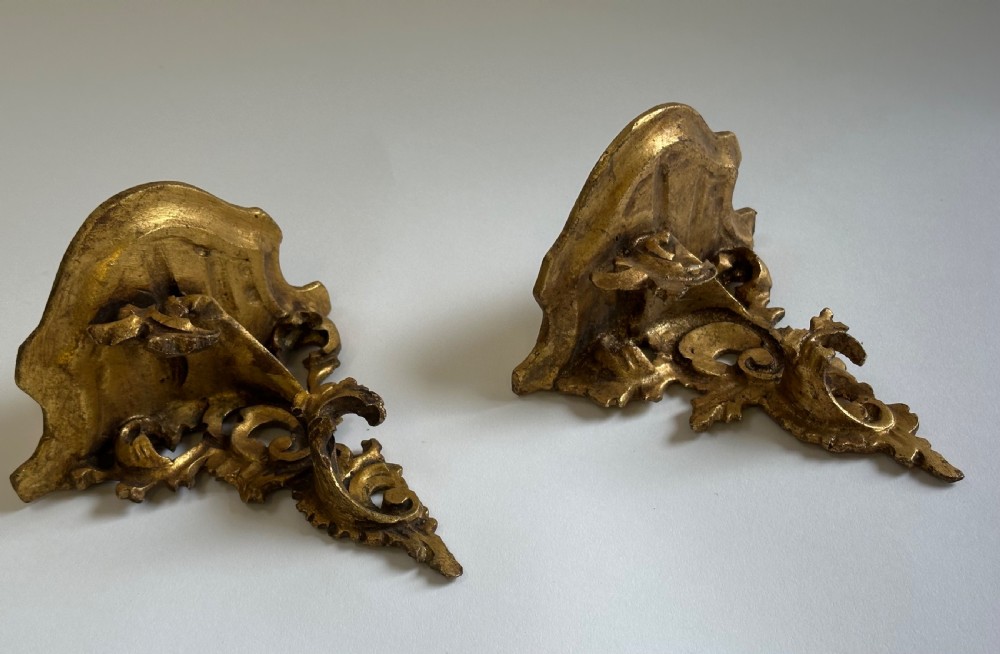 A pair of carved wood wall brackets with a hand-gilded gold finish featuring acanthus leaf scrollwork and flat top shelves. View VII