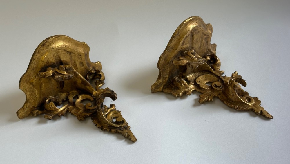 A pair of carved wood wall brackets with a hand-gilded gold finish featuring acanthus leaf scrollwork and flat top shelves. View XIV