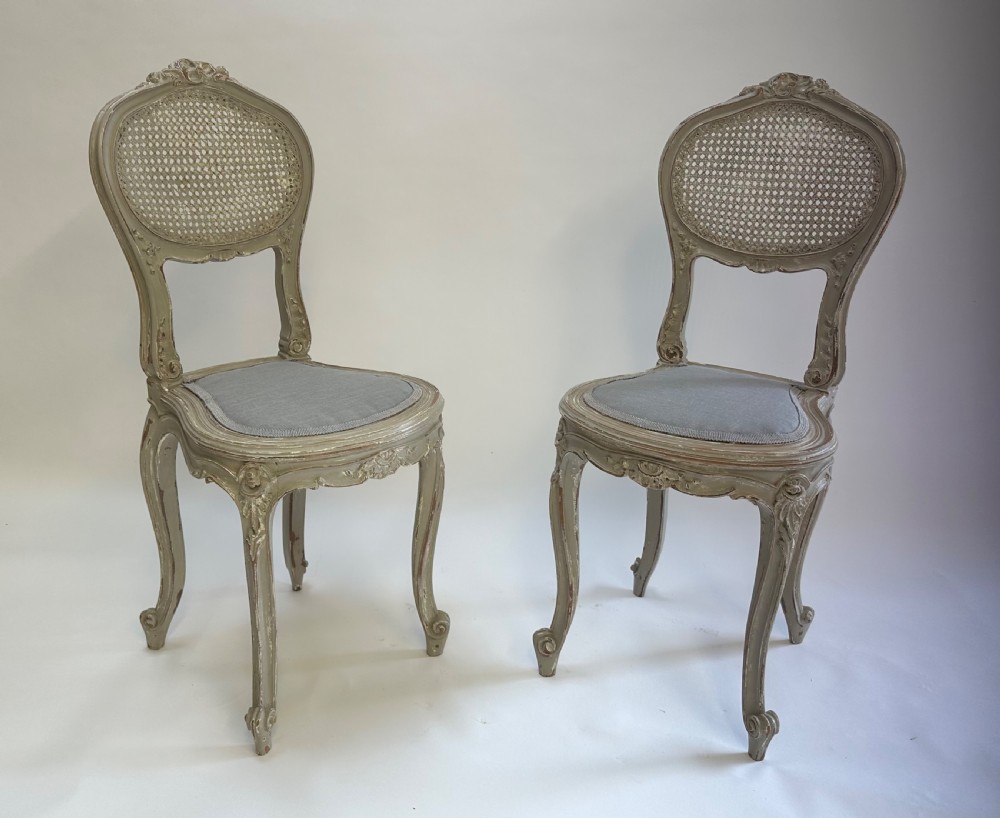 A pair of French Louis XV style side chairs with distressed grey painted frames, woven cane oval backrests, and grey fabric upholstered seats.