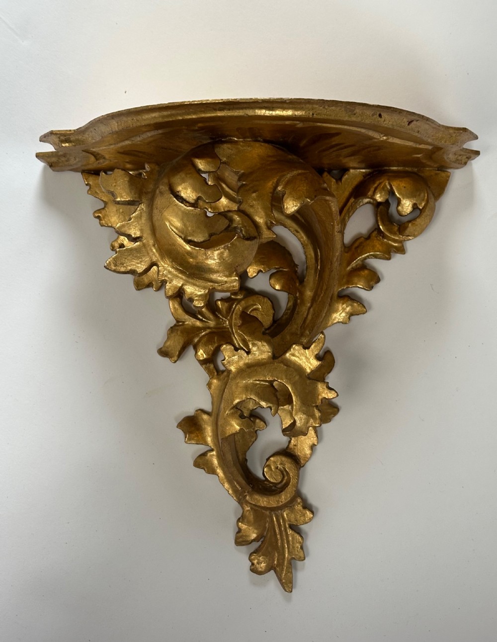 An Italian carved wood wall bracket featuring ornate acanthus leaf scrollwork with a gilded gold leaf finish supporting a semi-circular shelf top.
