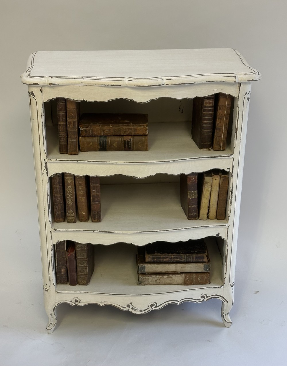 A small cream painted French open bookcase featuring scalloped shelf edges, panelled sides, and cabriole legs with distressed finish. View II