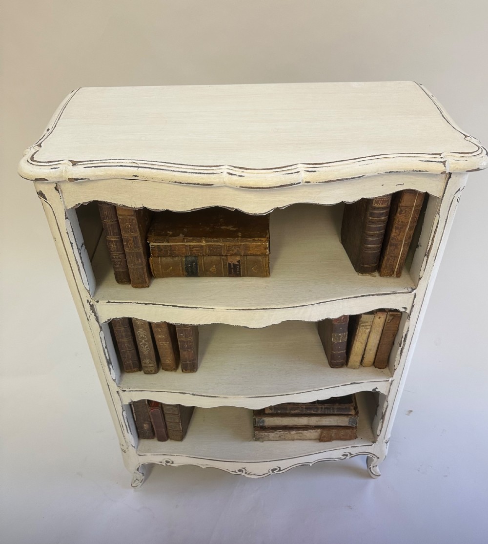 A small cream painted French open bookcase featuring scalloped shelf edges, panelled sides, and cabriole legs with distressed finish. View III
