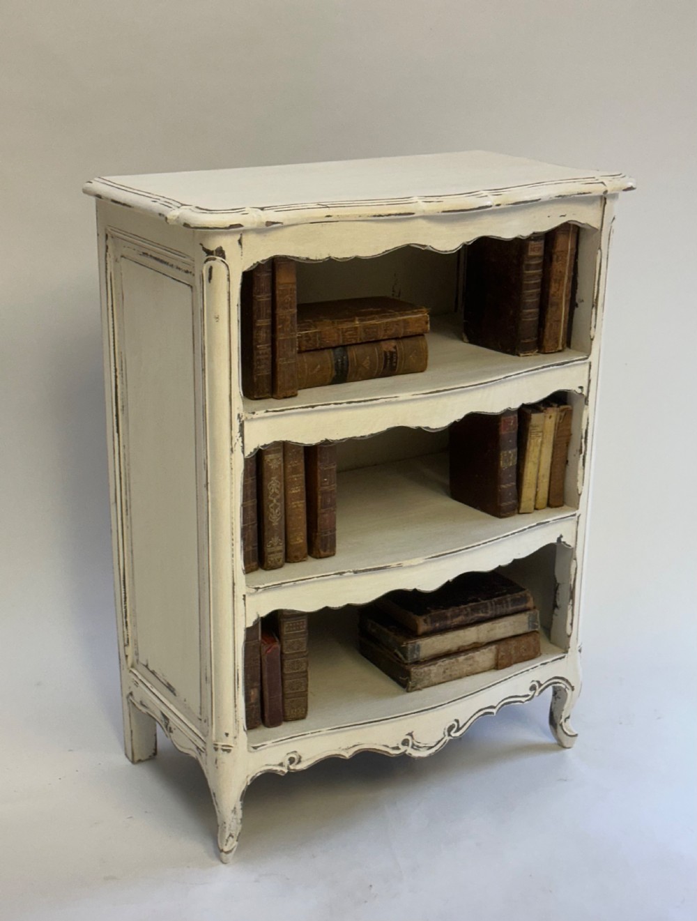 A small cream painted French open bookcase featuring scalloped shelf edges, panelled sides, and cabriole legs with distressed finish. View IV