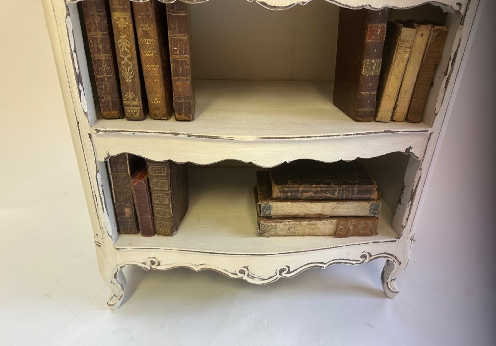 A small cream painted French open bookcase featuring scalloped shelf edges, panelled sides, and cabriole legs with distressed finish. View V