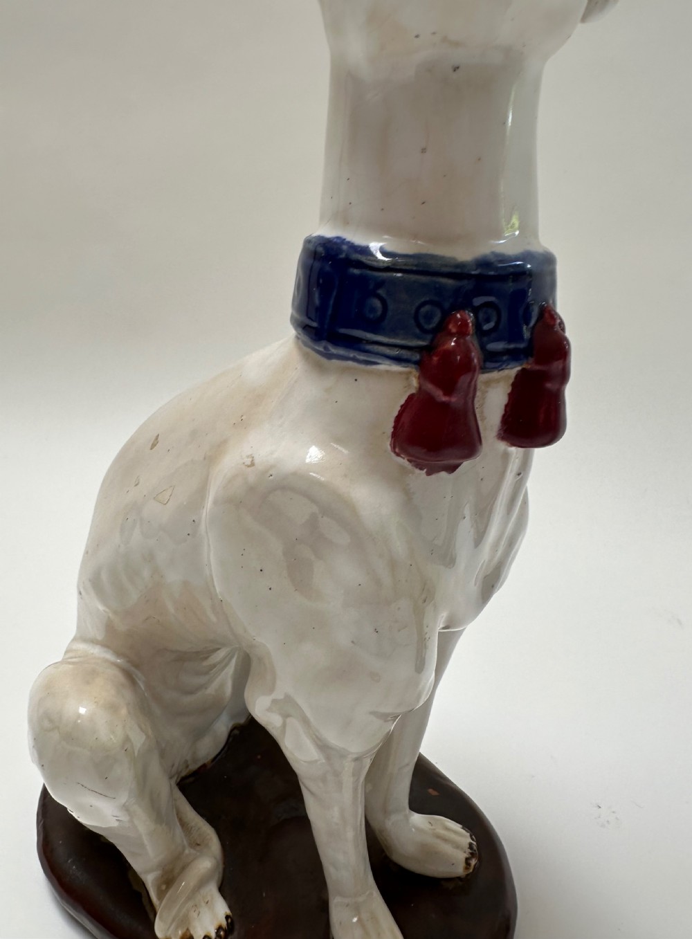 A pair of seated white ceramic greyhound sculptures with cobalt blue collars, red tassels, and brown oval bases. View II