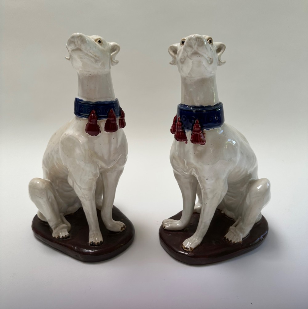 A pair of seated white ceramic greyhound sculptures with cobalt blue collars, red tassels, and brown oval bases. View III