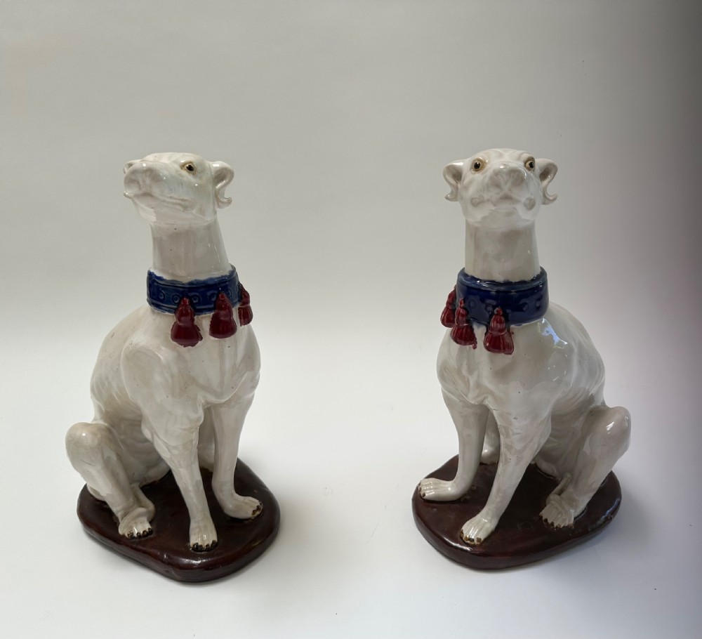 A pair of seated white ceramic greyhound sculptures with cobalt blue collars, red tassels, and brown oval bases. View IV