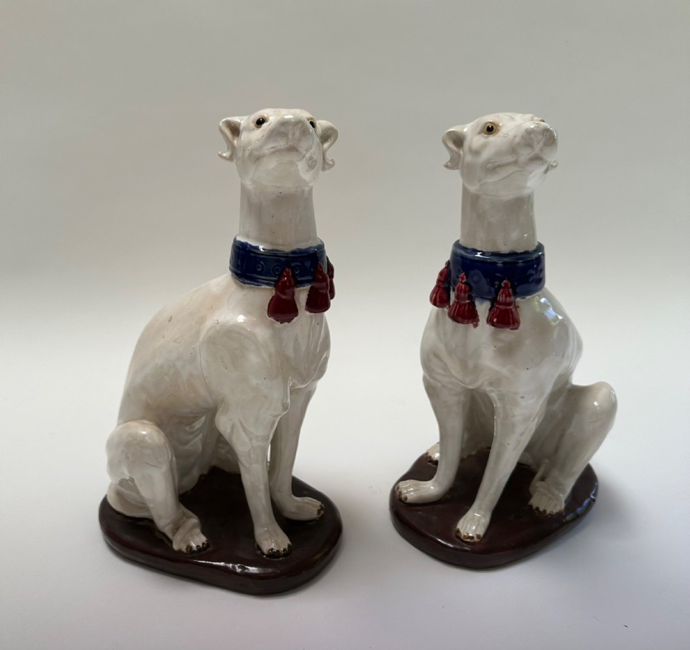 A pair of seated white ceramic greyhound sculptures with cobalt blue collars, red tassels, and brown oval bases. View V