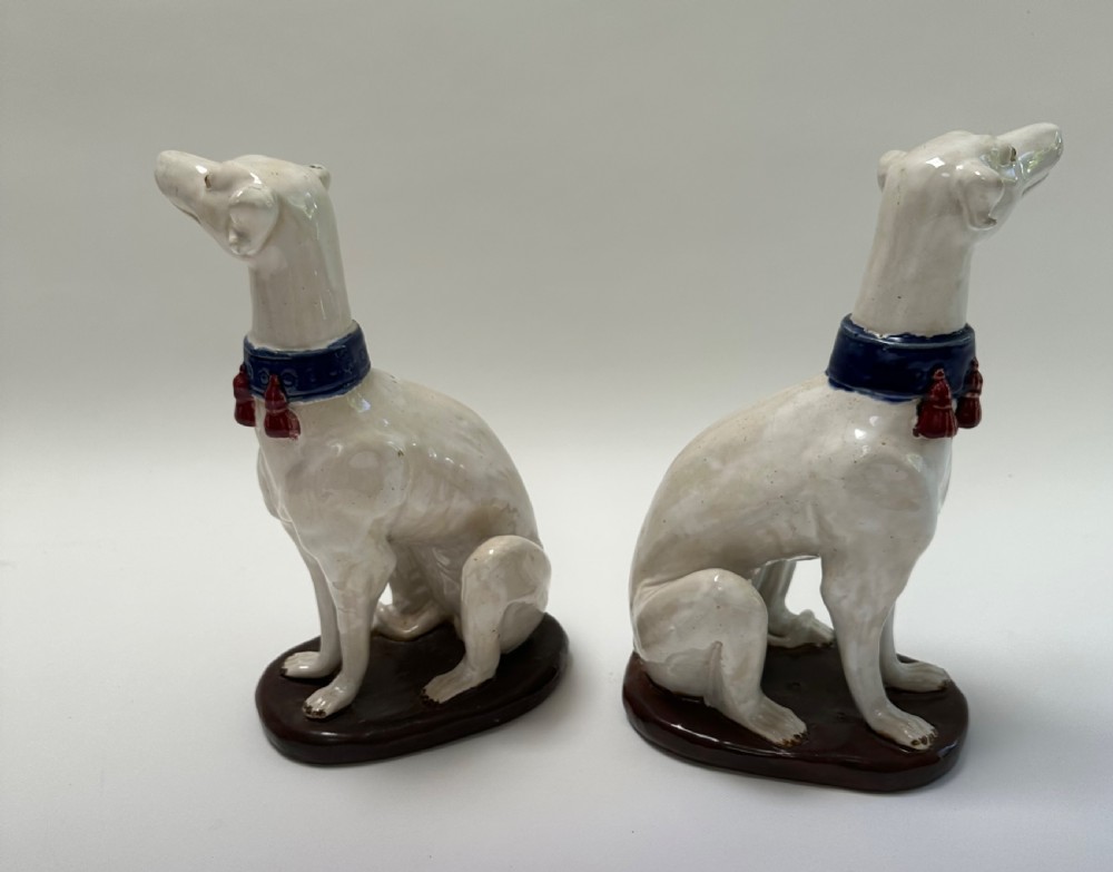 A pair of seated white ceramic greyhound sculptures with cobalt blue collars, red tassels, and brown oval bases. View VI