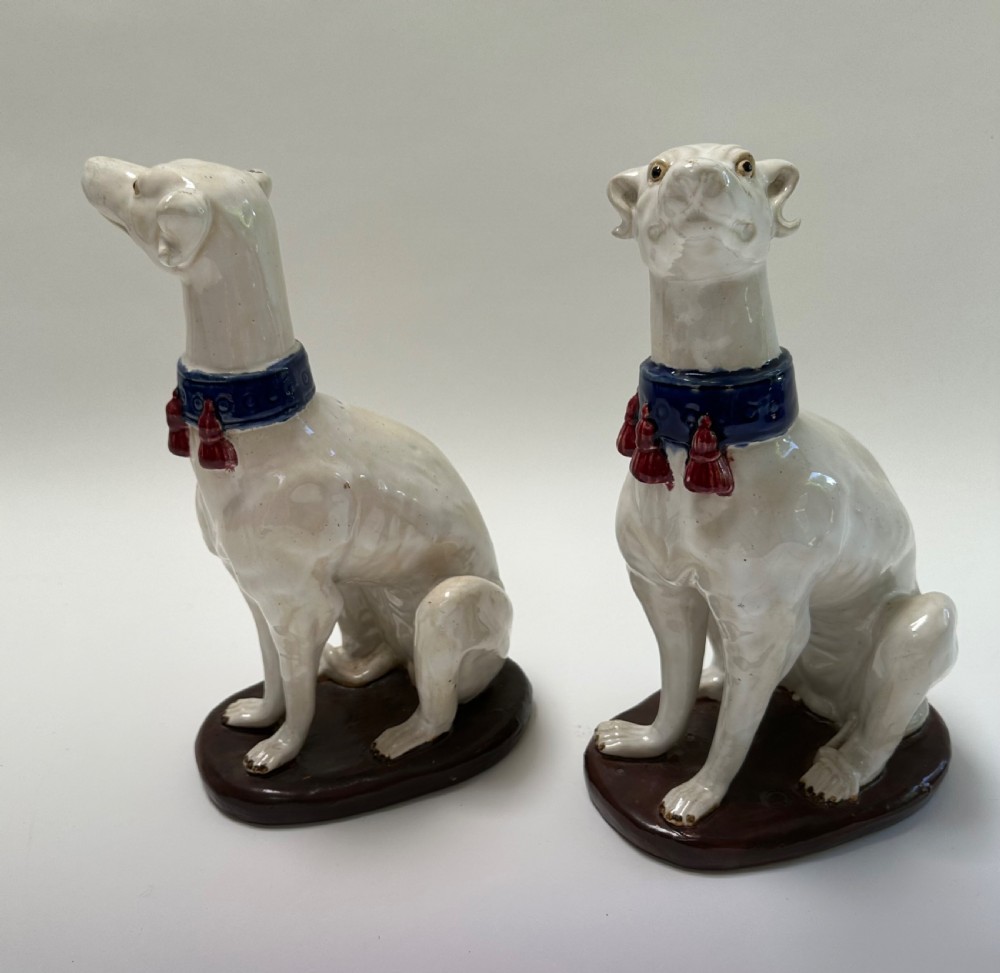 A pair of seated white ceramic greyhound sculptures with cobalt blue collars, red tassels, and brown oval bases. View VII