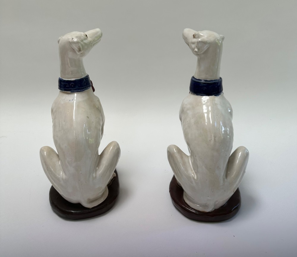 A pair of seated white ceramic greyhound sculptures with cobalt blue collars, red tassels, and brown oval bases. View VIII
