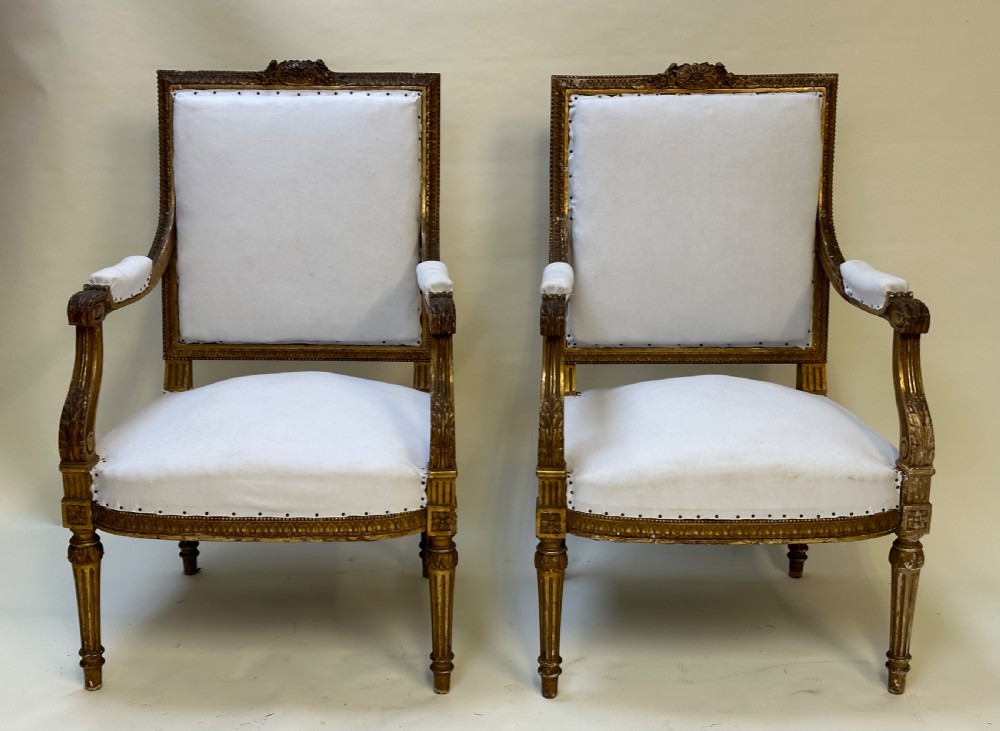 A pair of French Neoclassical giltwood armchairs featuring carved frames, fluted legs, and white fabric upholstery.