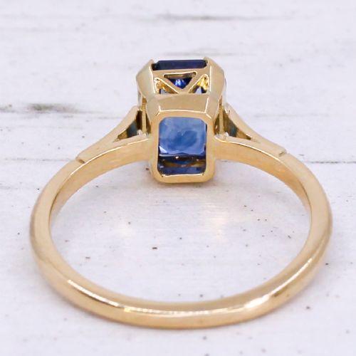 A yellow gold solitaire ring featuring an emerald-cut blue sapphire secured in a corner-claw setting. View V