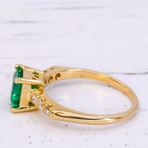 A yellow gold ring featuring a central square-cut green emerald held in a four-prong setting, accented by three small round diamonds on each shoulder. View IV