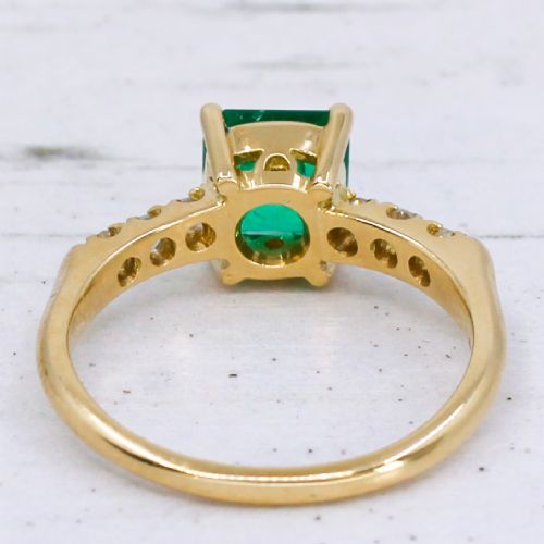 A yellow gold ring featuring a central square-cut green emerald held in a four-prong setting, accented by three small round diamonds on each shoulder. View V