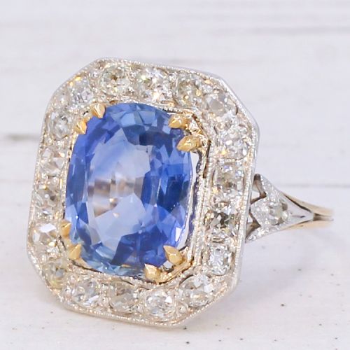 An antique Edwardian cluster ring featuring a central cushion-cut blue Ceylon sapphire surrounded by a halo of diamonds set in platinum and yellow gold.