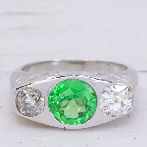 fig 2. A white metal ring featuring a central round green tsavorite garnet flanked by two old cut diamonds set within a carved band.