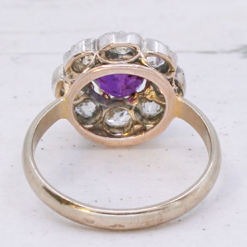 An antique cluster ring featuring a central oval purple-red ruby surrounded by ten old cut diamonds in a scalloped white metal setting. View V