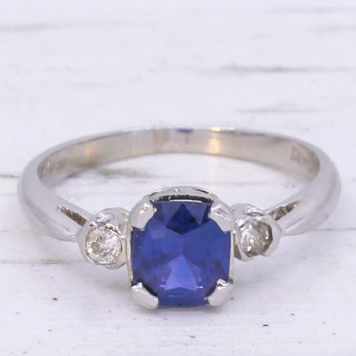 fig 2. A platinum ring featuring a central cushion-cut blue sapphire held in a claw setting, flanked by a small round diamond on each shoulder.