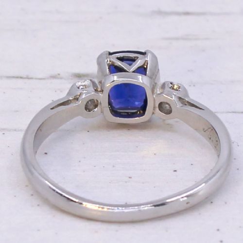 A platinum ring featuring a central cushion-cut blue sapphire held in a claw setting, flanked by a small round diamond on each shoulder. View VI