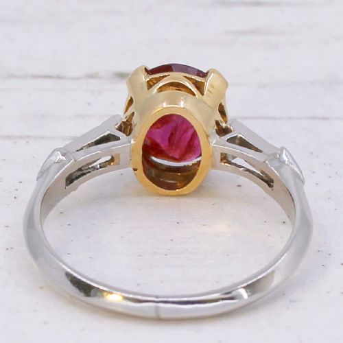 A vintage Art Deco solitaire ring featuring an oval red ruby in a gold setting on a silver-toned metal band with diamond-set shoulders. View V