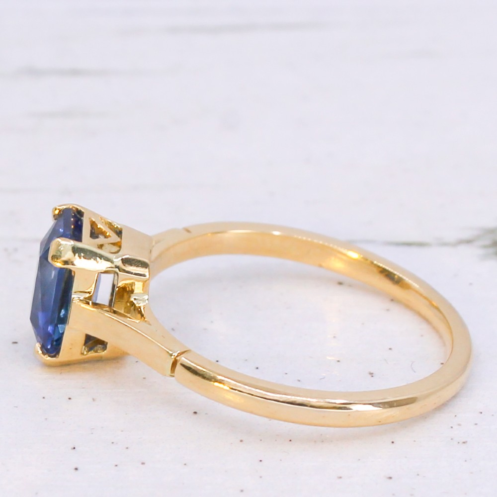 A yellow gold solitaire ring featuring an emerald-cut blue sapphire secured in a corner-claw setting. View IV
