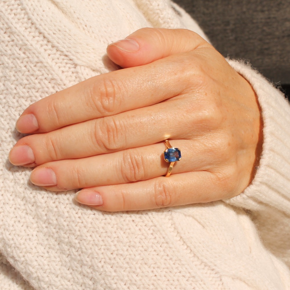 A yellow gold solitaire ring featuring an emerald-cut blue sapphire secured in a corner-claw setting. View VI