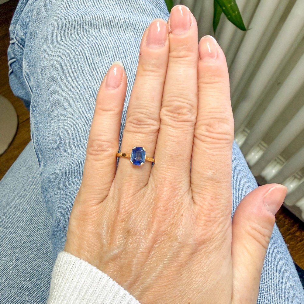 A yellow gold solitaire ring featuring an emerald-cut blue sapphire secured in a corner-claw setting. View VII