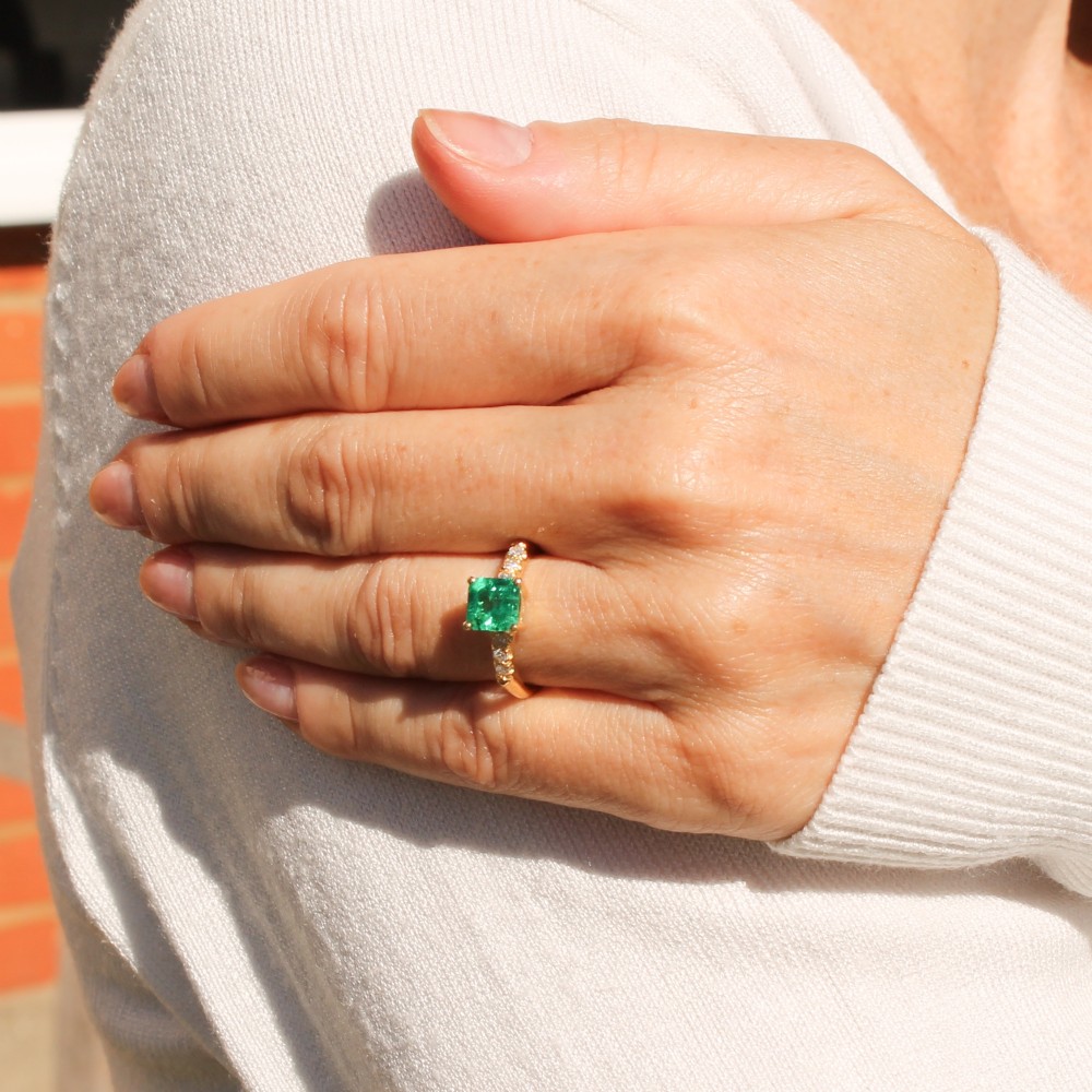 A yellow gold ring featuring a central square-cut green emerald held in a four-prong setting, accented by three small round diamonds on each shoulder. View VI