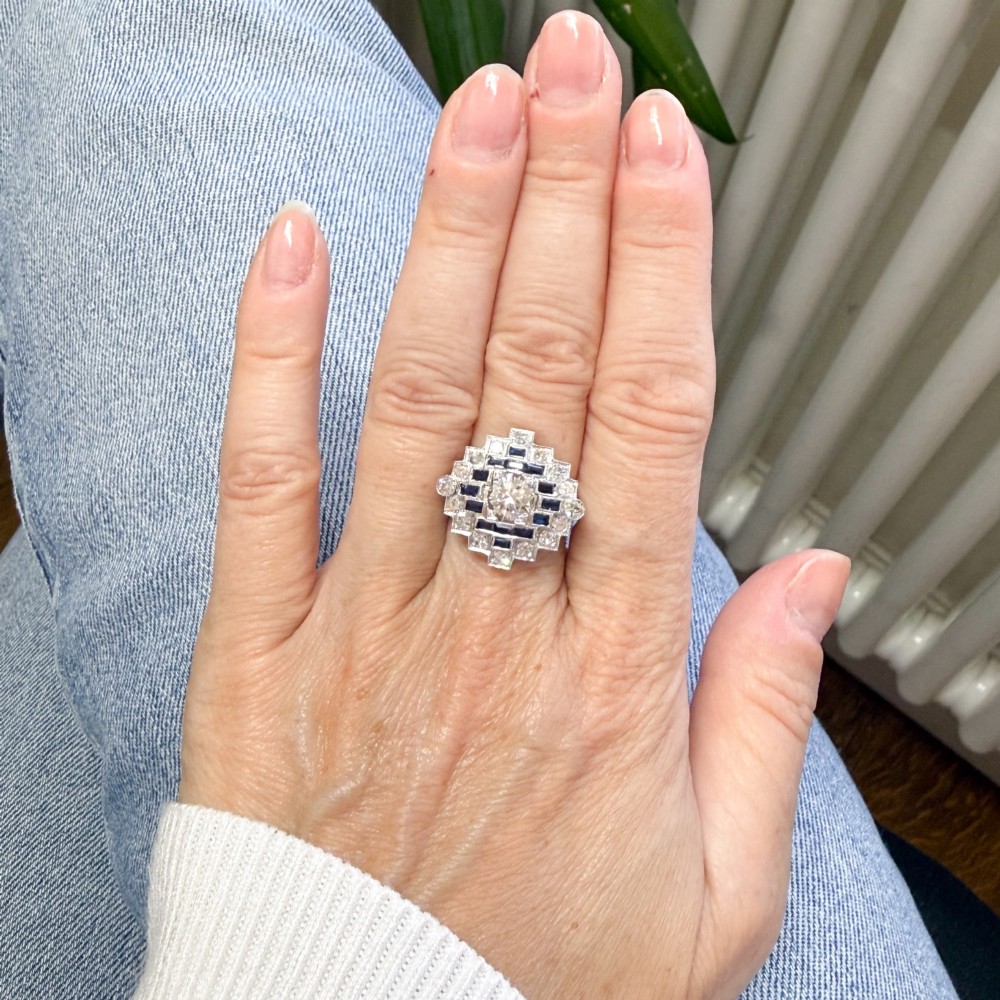 An Art Deco diamond and sapphire cluster ring featuring a central brilliant-cut diamond surrounded by geometric borders of blue sapphires and smaller diamonds. View VI