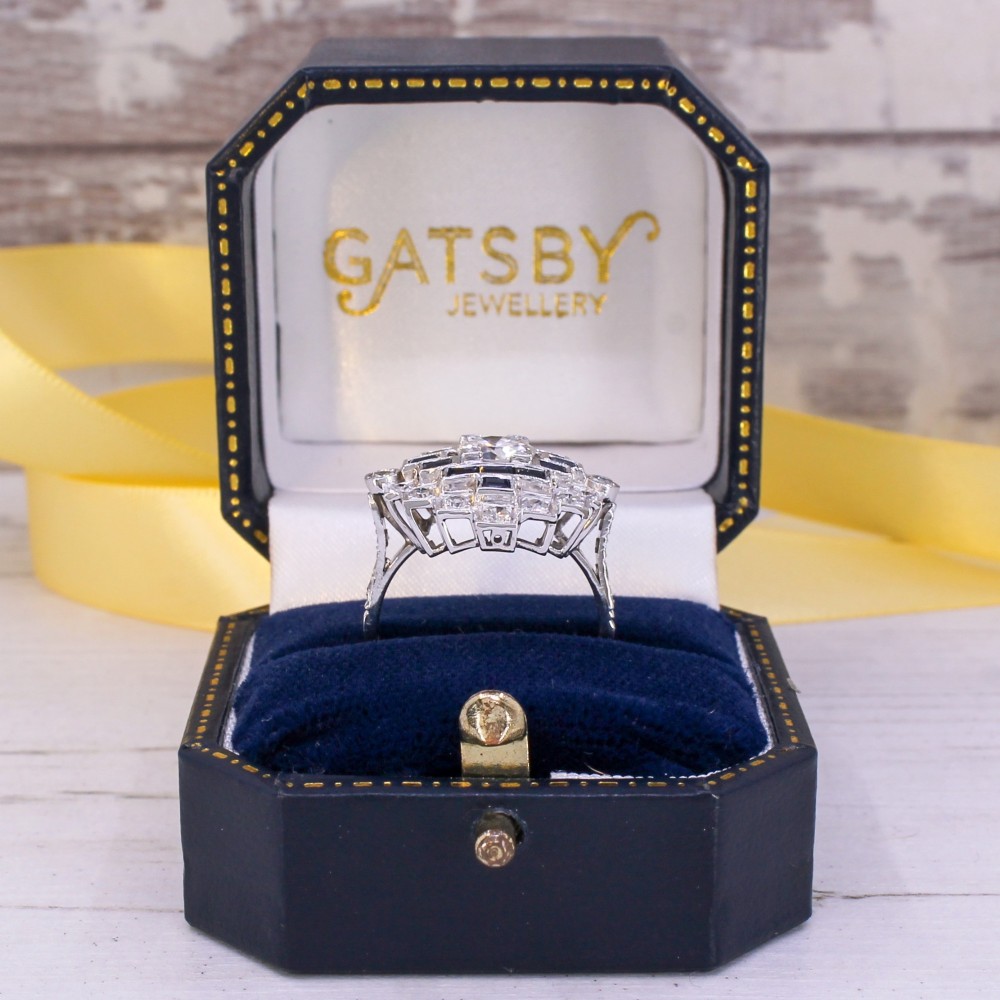 An Art Deco diamond and sapphire cluster ring featuring a central brilliant-cut diamond surrounded by geometric borders of blue sapphires and smaller diamonds. View IX