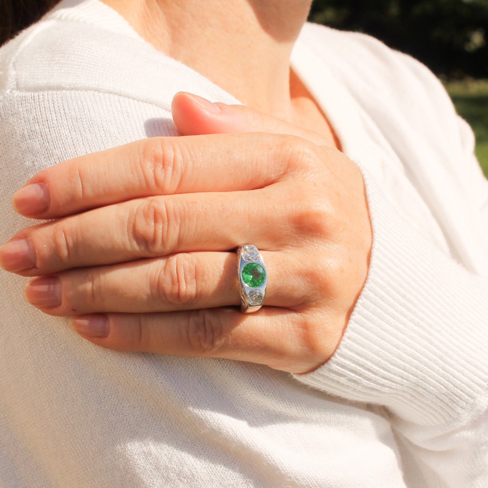 A white metal ring featuring a central round green tsavorite garnet flanked by two old cut diamonds set within a carved band. View II