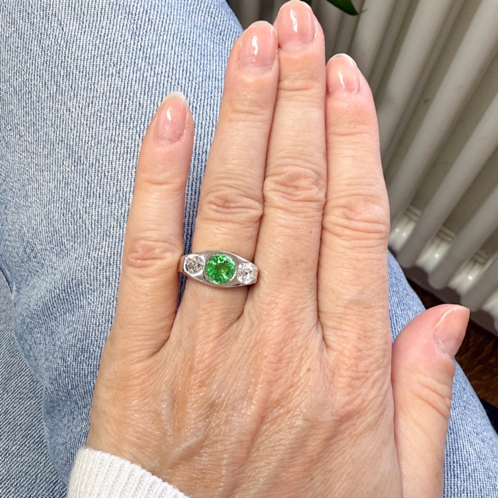 A white metal ring featuring a central round green tsavorite garnet flanked by two old cut diamonds set within a carved band. View VI