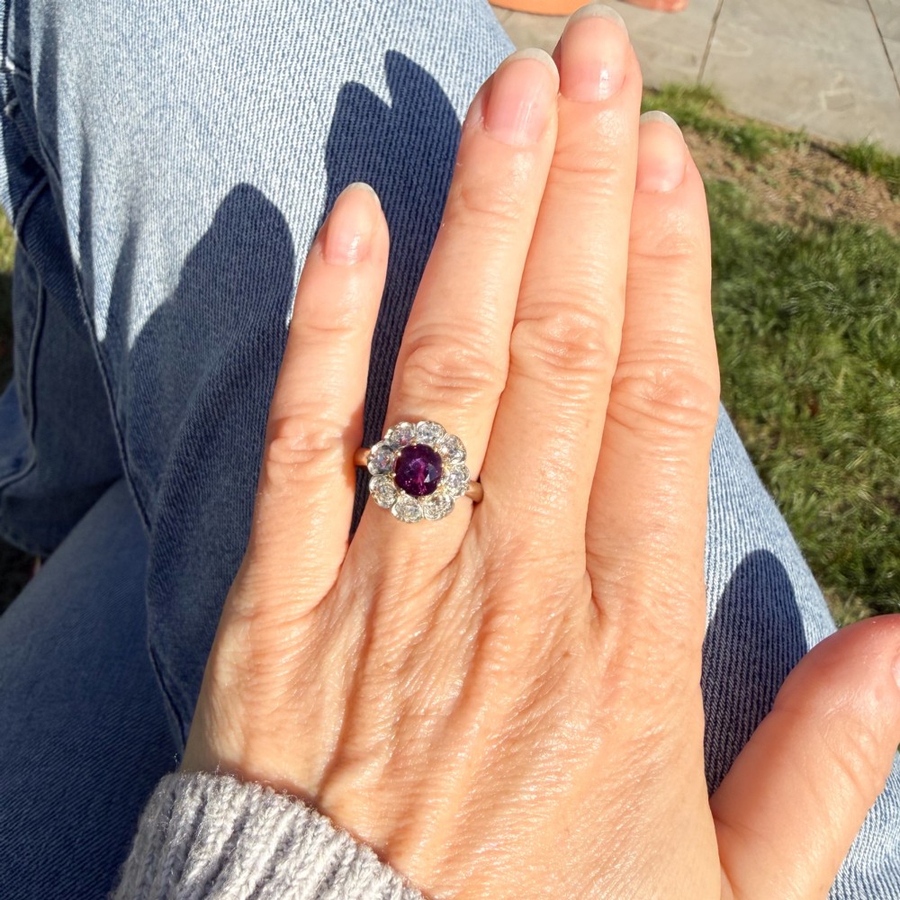 An antique cluster ring featuring a central oval purple-red ruby surrounded by ten old cut diamonds in a scalloped white metal setting. View VI