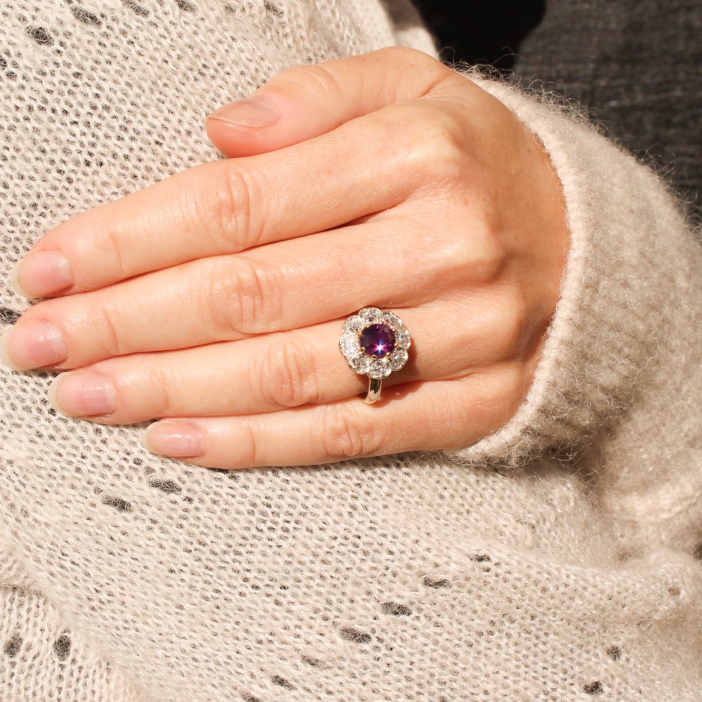 An antique cluster ring featuring a central oval purple-red ruby surrounded by ten old cut diamonds in a scalloped white metal setting. View VII