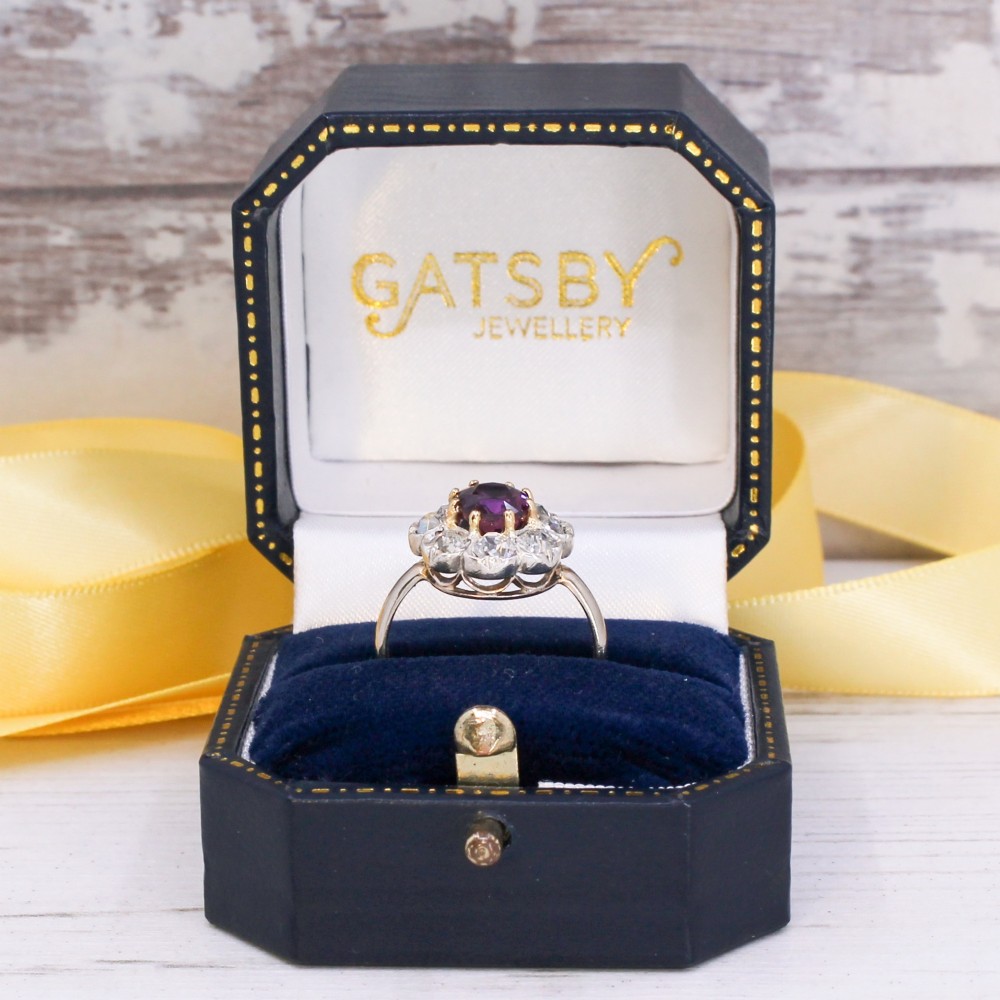 An antique cluster ring featuring a central oval purple-red ruby surrounded by ten old cut diamonds in a scalloped white metal setting. View IX