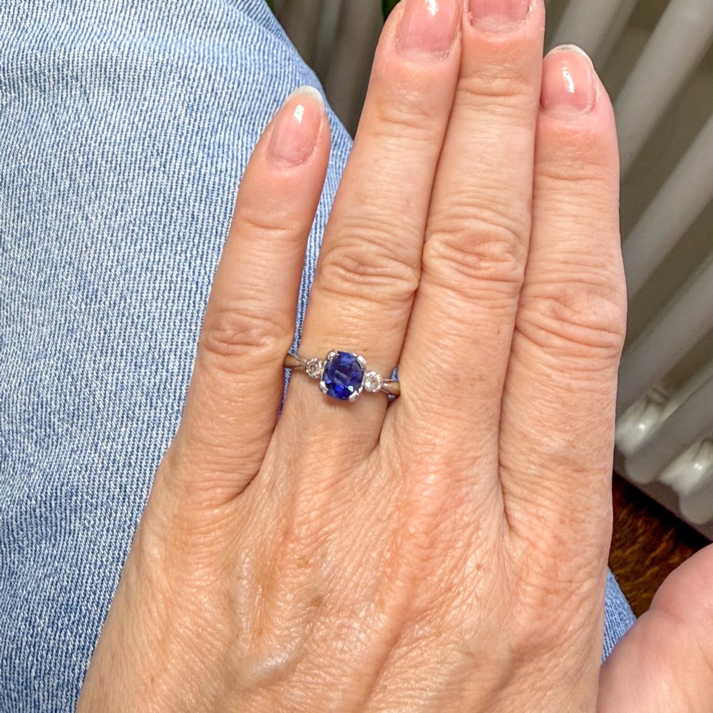 A platinum ring featuring a central cushion-cut blue sapphire held in a claw setting, flanked by a small round diamond on each shoulder. View VII