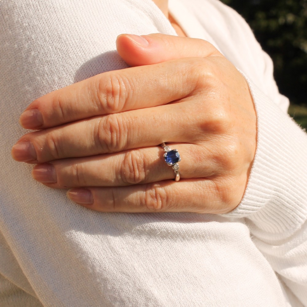 A platinum ring featuring a central cushion-cut blue sapphire held in a claw setting, flanked by a small round diamond on each shoulder. View VIII
