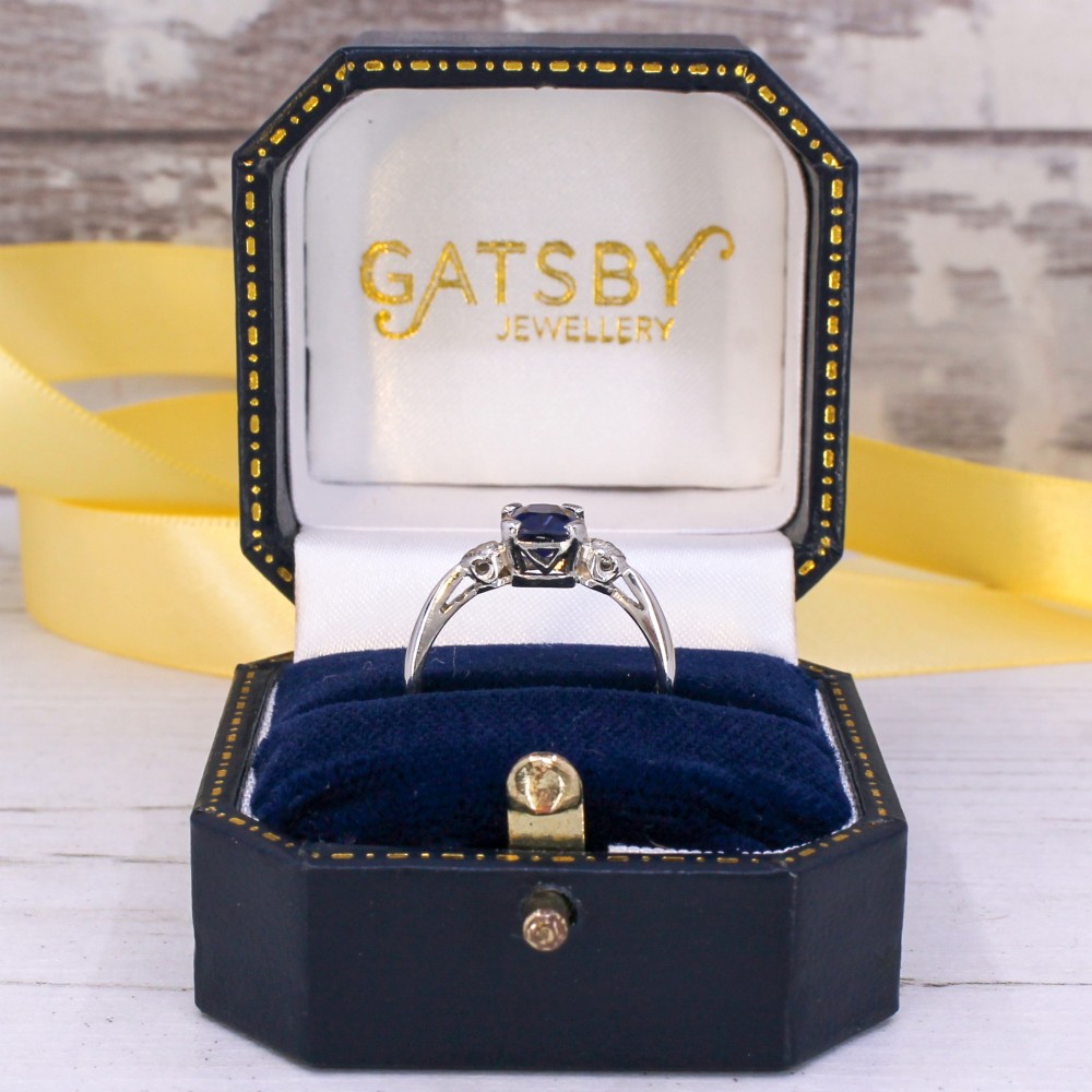 A platinum ring featuring a central cushion-cut blue sapphire held in a claw setting, flanked by a small round diamond on each shoulder. View X