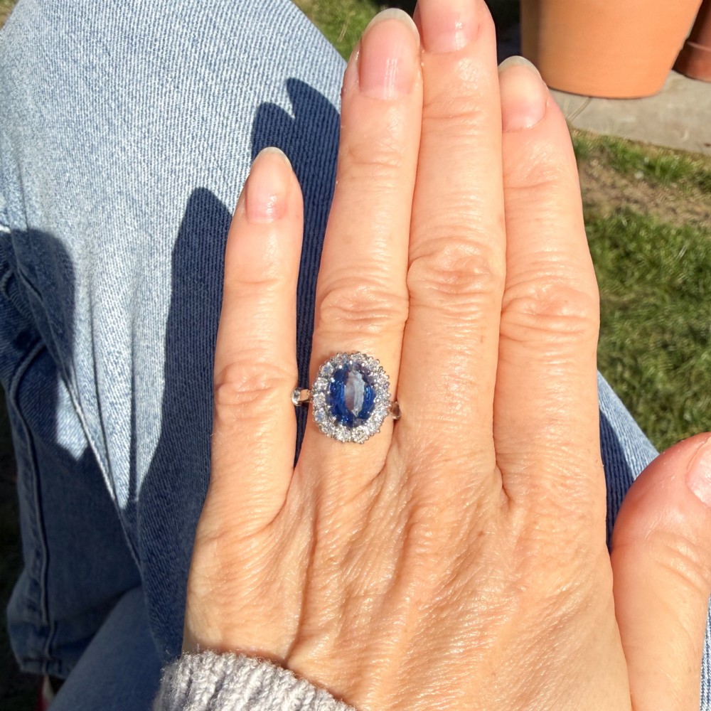 An oval blue Ceylon sapphire encircled by a halo of brilliant diamonds set on a silver metal band. View VI