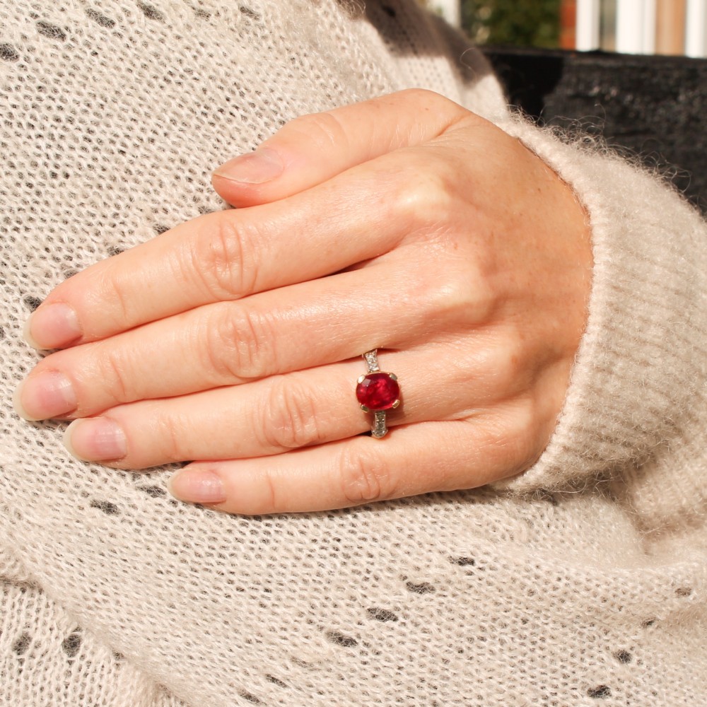 A vintage Art Deco solitaire ring featuring an oval red ruby in a gold setting on a silver-toned metal band with diamond-set shoulders. View II