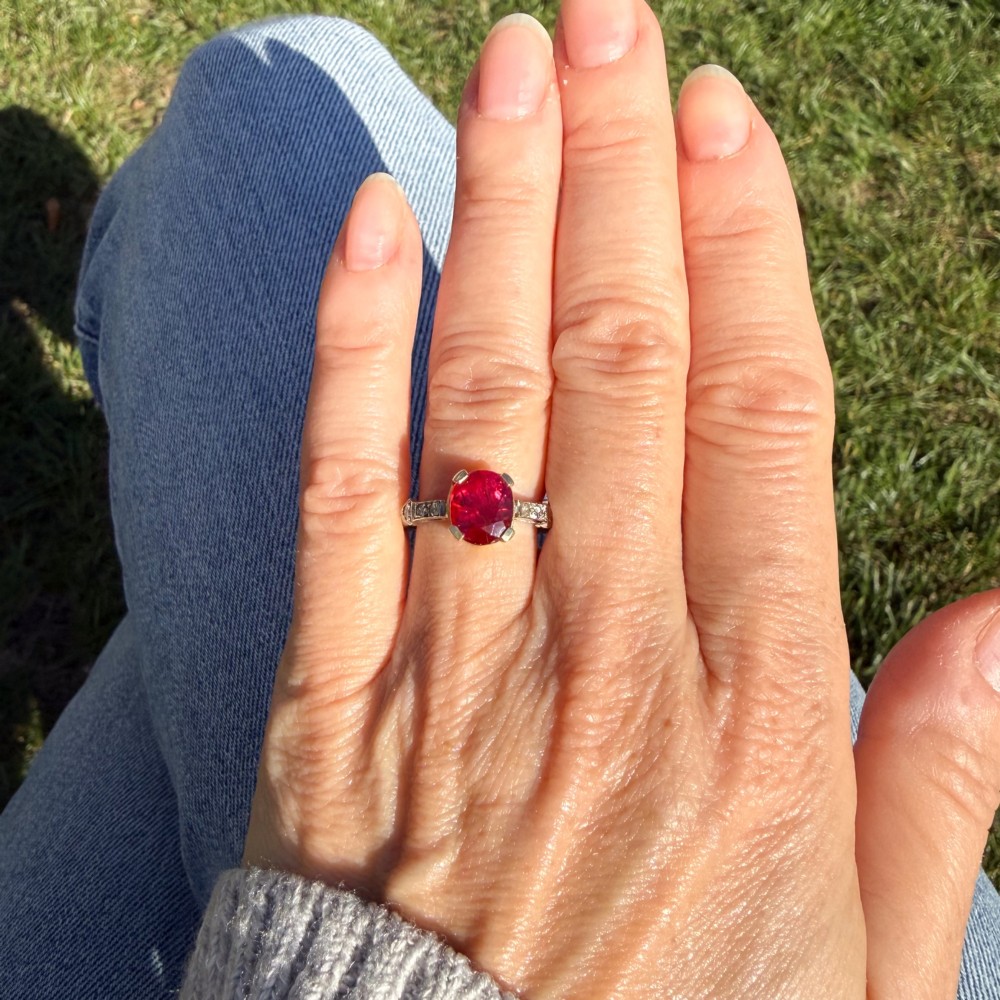 A vintage Art Deco solitaire ring featuring an oval red ruby in a gold setting on a silver-toned metal band with diamond-set shoulders. View VI