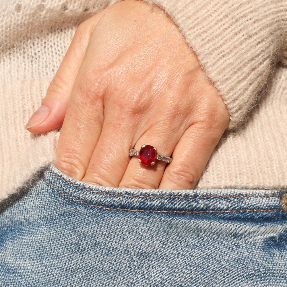 A vintage Art Deco solitaire ring featuring an oval red ruby in a gold setting on a silver-toned metal band with diamond-set shoulders. View VII