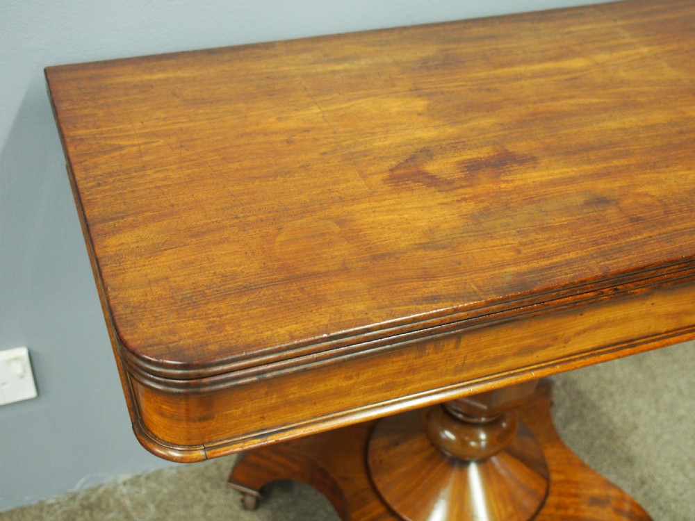 A 19th-century William IV mahogany fold-over table featuring a rounded rectangular top, a thick turned central pedestal, a concave quadform base, and brass castors. View V