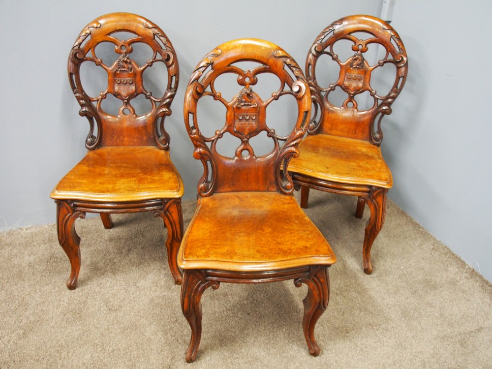 Set of three Victorian mahogany hall chairs featuring openwork carved backs inset with central heraldic crests and shaped front legs.